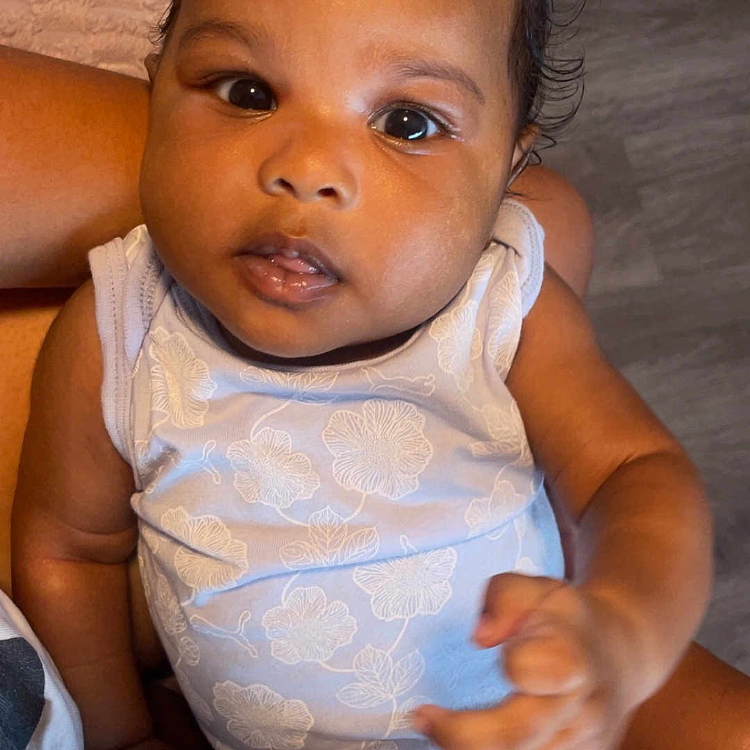 D Aria is registered to the contest to win money with this photo: arm, baby, child, closeup, curious, cute, expression, face, floor, floral_pattern, hair_bow, hand, indoor, infant, onesie, person, portrait, sitting, skin, young