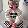 baby, beanie, bed, child, clothing, cozy, cross_legged, cute, face, happy, indoor, person, playful, portrait, smiling, socks, striped_pants, sweater, wall_decal, young_child