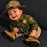 baby, badge, binoculars, boots, child, costume, couch, cute, green_shirt, happy, hat, indoors, park_ranger, person, portrait, sitting, smiling, toddler, uniform, young_child