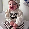 baby, beanie, bed, child, clothing, cozy, cross_legged, cute, face, happy, indoor, person, playful, portrait, smiling, socks, striped_pants, sweater, wall_decal, young_child
