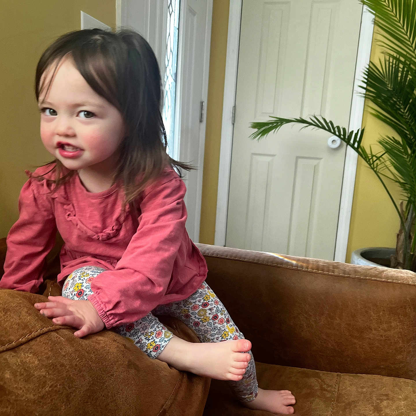 Corinne joined the competition — help win amazing prizes! barefoot, brown_couch, child, couch, door, face, floral_leggings, foot, hair, hand, home, indoor, living_room, pink_clothing, plant, playful, portrait, smiling, toddler, yellow_wall