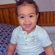 Elora is registered to the contest to win money with this photo: toddler, child, smiling, happy, indoor, bed, clothing, white_shirt, dark_pants, curly_hair, wood_paneling, blanket, face, person, cute, young_child, portrait, sitting, joyful, expression