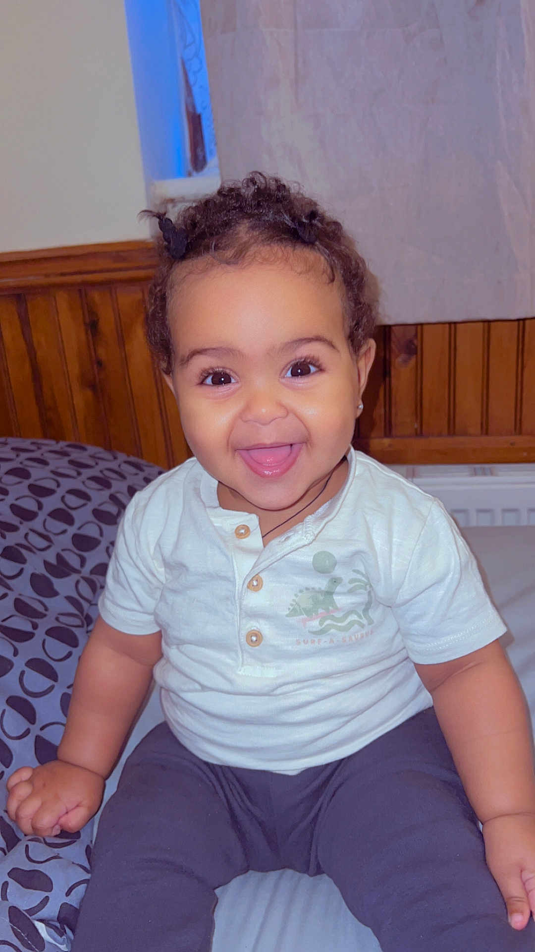 Elora is registered to the contest to win money with this photo: toddler, child, smiling, happy, indoor, bed, clothing, white_shirt, dark_pants, curly_hair, wood_paneling, blanket, face, person, cute, young_child, portrait, sitting, joyful, expression