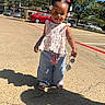 blue_pants, braided_hair, cars, child, outdoor, parking_lot, pavement, person, playful, red_bows, sandals, shadow, smile, sunglasses, sunlight, sunny, toddler, trees, white_flower_clips, white_top