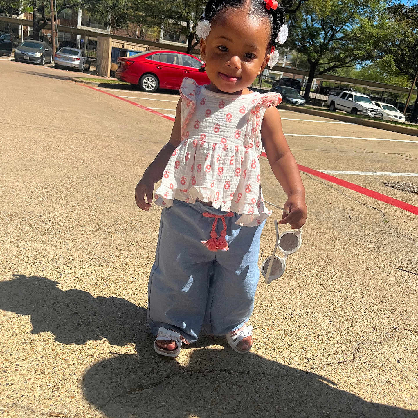 Ahbri is registered to the contest to win money with this photo: blue_pants, braided_hair, cars, child, outdoor, parking_lot, pavement, person, playful, red_bows, sandals, shadow, smile, sunglasses, sunlight, sunny, toddler, trees, white_flower_clips, white_top