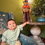 Enzo is registered to the contest to win money with this photo: armchair, blue_socks, casual_clothing, child, cozy, decor, figurine, green_wall, home, indoor, patterned_fabric, plant, pottery, reaching, shirt, shorts, smile, table, toddler, toy