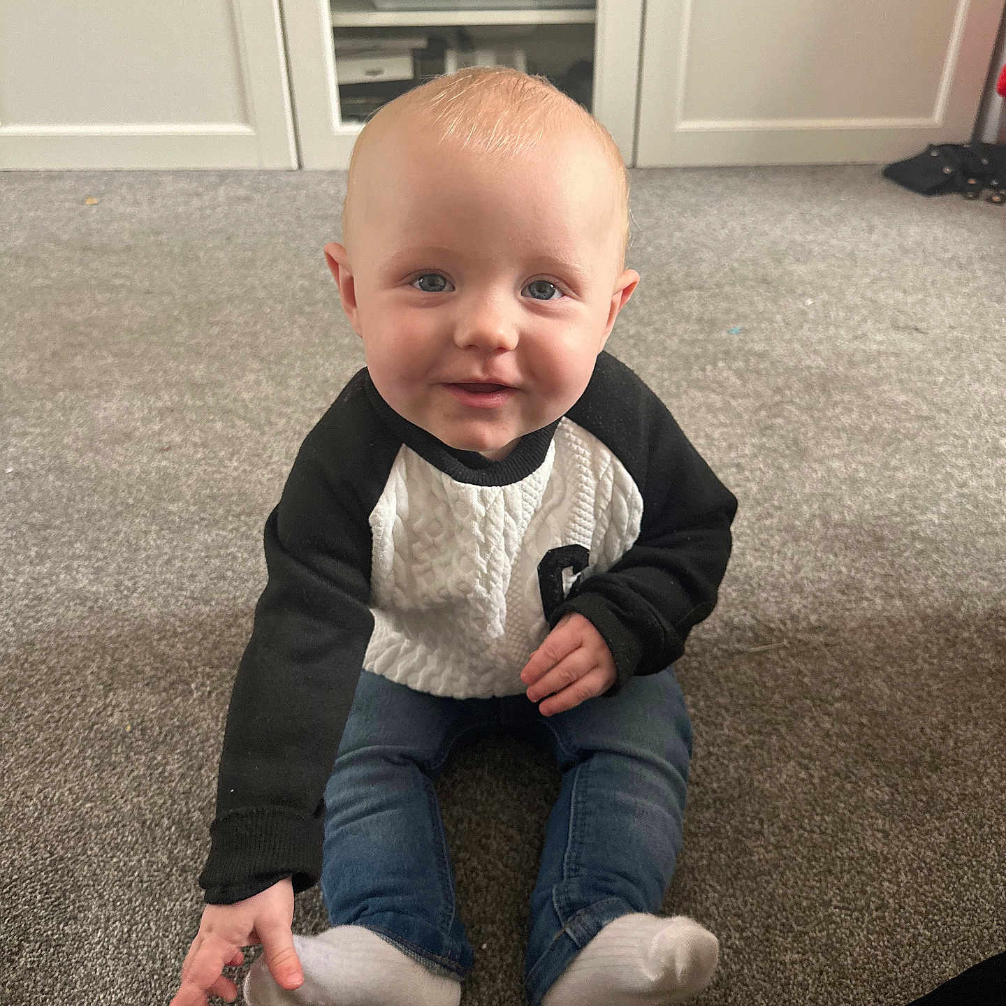 Louie is registered to the contest to win money with this photo: baby, blue_eyes, cabinet, carpet, child, cute, floor, furniture, home, indoor, infant, jeans, light_hair, person, portrait, shelf, sitting, smiling, socks, sweater