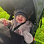 baby, child, smiling, stroller, hat, pom_poms, coat, fuzzy_clothing, outdoor, green_grass, sunlight, happy, infant, seatbelt, baby_stroller, cute, warm_clothing, portrait, young_child, daytime