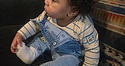 Arion is registered to the contest to win money with this photo: toddler, child, curly_hair, denim_overalls, striped_shirt, socks, couch, blanket, indoor, furniture, baby, seated, side_profile, cute, curious, hand, foot, cozy, casual_clothing, home