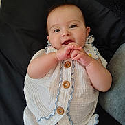 Rhiannon is registered to the contest to win money with this photo: baby, infant, smile, hands_clasped, white_romper, buttons, ruffle, cheeks, eyes, ear, portrait, lying_down, pillow, black_background, cute, skin, person, indoor, playful, closeup