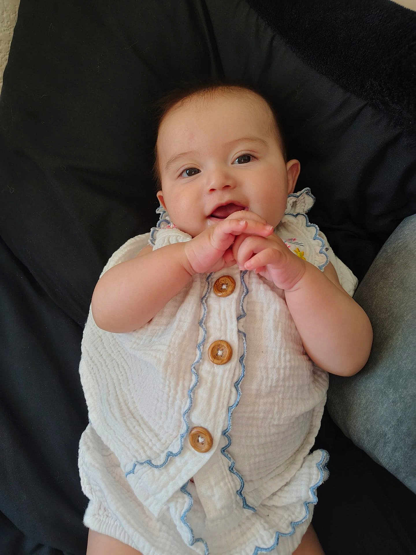Rhiannon is registered to the contest to win money with this photo: baby, infant, smile, hands_clasped, white_romper, buttons, ruffle, cheeks, eyes, ear, portrait, lying_down, pillow, black_background, cute, skin, person, indoor, playful, closeup
