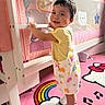 Daniela is registered to the contest to win money with this photo: toddler, child, smiling, indoor, crib, bedroom, pink, colorful_clothing, overalls, sneakers, toy, rug, hello_kitty, rainbow, musical_notes, furniture, baby_room, cute, person, playful