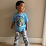 child, boy, standing, blue_tshirt, cartoon_characters, jeans, shoes, indoor, wooden_floor, plain_wall, curly_hair, smiling, casual_clothing, portrait, fashion, cute, confident, playful, person, floor
