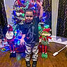 child, boy, christmas_tree, holiday_decorations, santa_doll, elf_doll, indoor, wooden_floor, festive_lights, pose, smile, black_shoes, pants, tshirt, decorations, toys, curtains, wallpaper, home, celebration