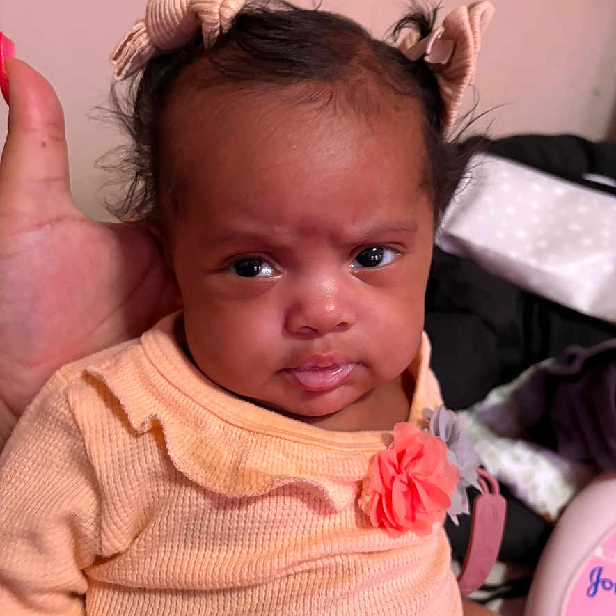 Lyliah joined the competition — help win amazing prizes! baby, child, closeup, cute, expression, face, flower_accessory, girl, hair_bow, hand, head_support, indoor, infant, peach_clothing, person, portrait, sitting, skin, soft_lighting, young