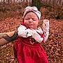 baby, infant, child, headband, red_dress, long_sleeve, patterned_clothing, puckered_face, closed_eyes, arm, tattoo, hand, outdoors, forest, fallen_leaves, autumn, tree, portrait, person_holding, cute_expression