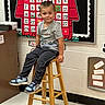child, boy, smile, stool, classroom, calendar, september, wall, shoes, pants, tshirt, hair, floor, furniture, education, indoor, young, cute, happy, sitting