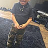 Kaiden is registered to the contest to win money with this photo: toddler, child, smiling, black_polo_shirt, camo_pants, standing, black_shoes, carpet, living_room, television, wooden_floor, smile, happy, earrings, necklace, cross_pendant, furniture, home_interior, person, indoor