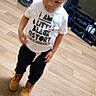 child, boy, cornrows, white_tshirt, boots, wooden_floor, indoor, standing, looking_down, casual_clothing, black_history, hair_style, young, person, flooring, home, fashion, cute, toddler, expression