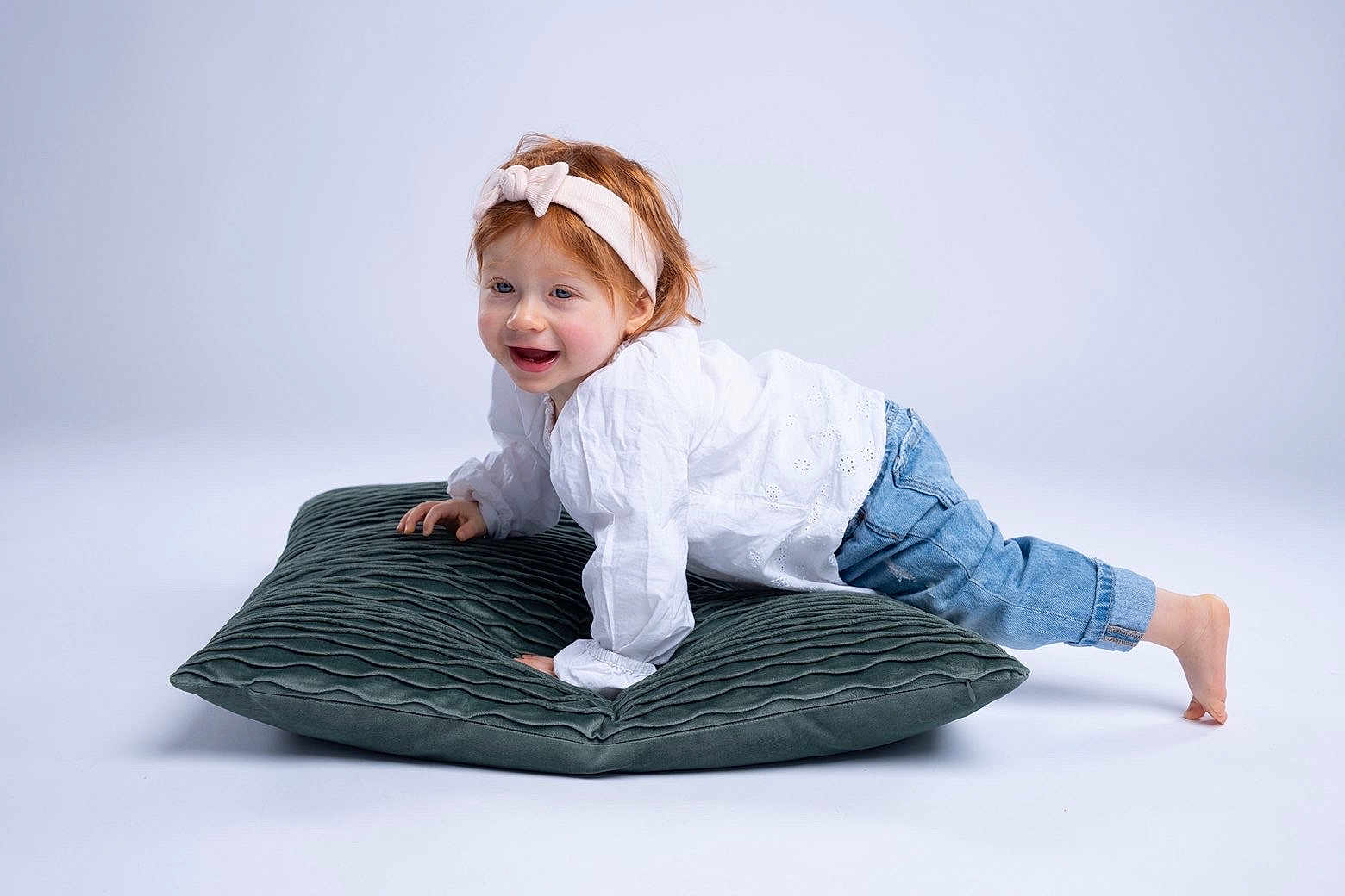 Inaya a rejoint le concours — aidez-le/la à gagner de superbes lots ! toddler, child, headband, red_hair, white_shirt, blue_jeans, cushion, smiling, playful, barefoot, studio, portrait, happy, crawling, indoors, cute, young_child, fashion, casual_wear, white_background