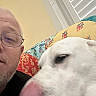 man, glasses, dog, white_dog, close_up, selfie, indoor, quilt, colorful_quilt, face, pet, animal, beard, home, window_blinds, nose, licking, casual, portrait, candid