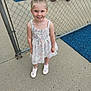 Hermonie is registered to the contest to win money with this photo: child, girl, smiling, pigtails, blonde_hair, blue_eyes, dress, floral_pattern, sneakers, pink_hearts, chain_link_fence, playground, concrete, outdoor, happy, person, young_child, standing, casual_clothing, daylight