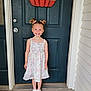 child, girl, smile, door, welcome_sign, pumpkin, hair_bows, dress, floral_pattern, sneakers, white_shoes, pink_hearts, front_porch, concrete_floor, blue_door, happy, standing, outdoor, person, home_entrance