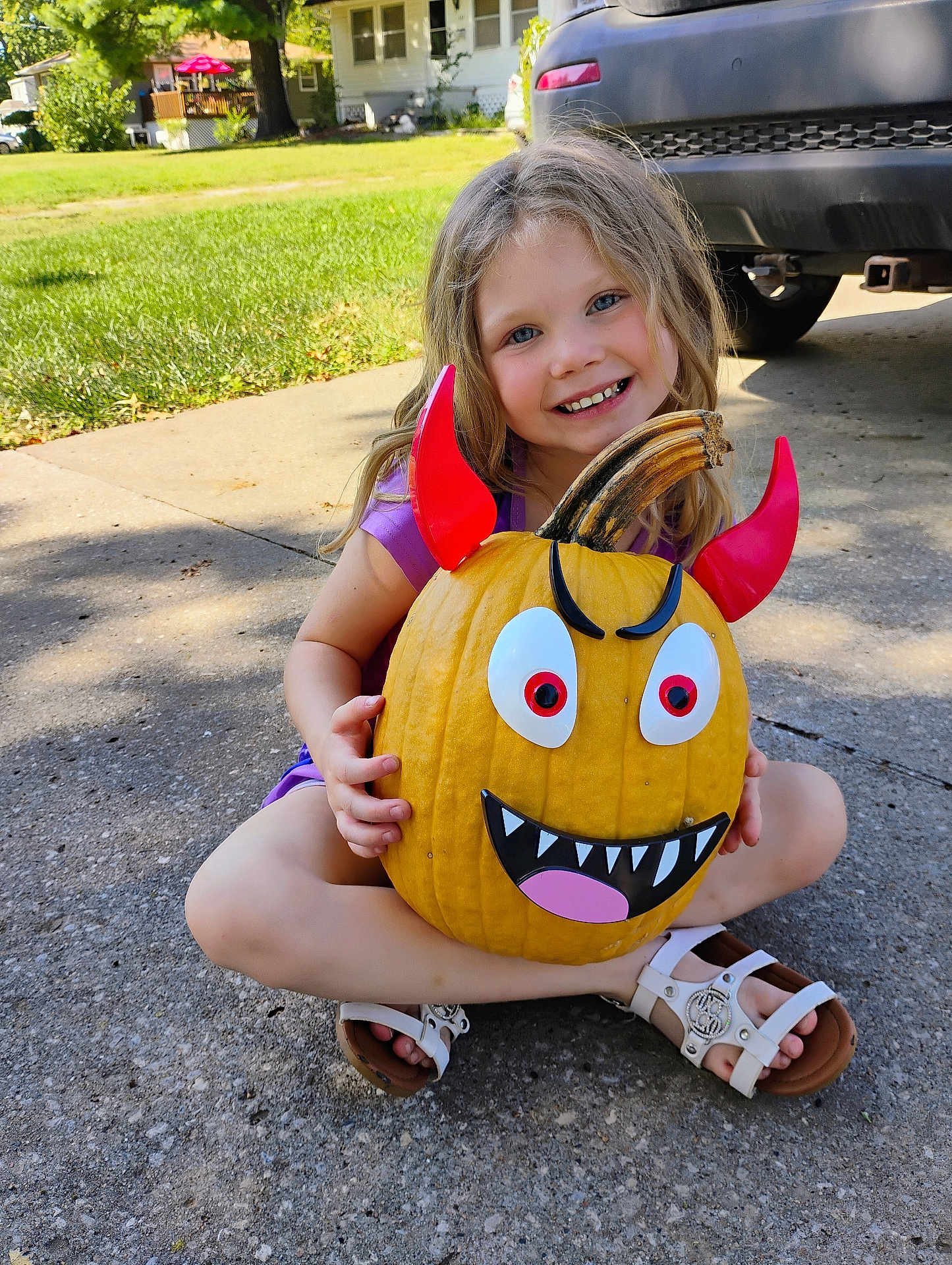 Hermonie is registered to the contest to win money with this photo: child, girl, pumpkin, devil_horns, smiling, outdoor, driveway, grass, house, car, daylight, purple_shirt, white_sandals, concrete, fall, holiday, decoration, happy, cute, playful