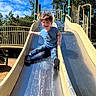 child, boy, slide, playground, outdoor, smiling, happy, blue_shirt, jeans, sneakers, metal_slide, sunny, daytime, trees, fence, play_equipment, nature, fun, leisure, park