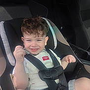 Elijah is registered to the contest to win money with this photo: child, toddler, car_seat, safety_harness, smiling, curly_hair, seatbelt, car_interior, sunlight, happy, young_child, casual_clothing, striped_shorts, white_shirt, portrait, infant, baby, seat, vehicle, cute