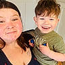 background_wall, brown_hair, casual_clothing, child, face, family, green_shirt, happy, holding, indoor, light_skin, navy_top, person, portrait, short_hair, smile, tattoo, toy, woman, young_child