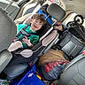 child, car_seat, smiling, green_shirt, car_interior, seatbelt, headrest, luggage, stroller, toy, window, parking_lot, happy, person, seat, bag, storage, clothing, baby, travel