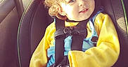 Zayden is registered to the contest to win money with this photo: toddler, child, car_seat, safety_harness, curly_hair, red_bow, yellow_sleeves, blue_overall, smiling, portrait, indoors, car_interior, seatbelt, cute, young_child, happy, person, baby, clothing, seat