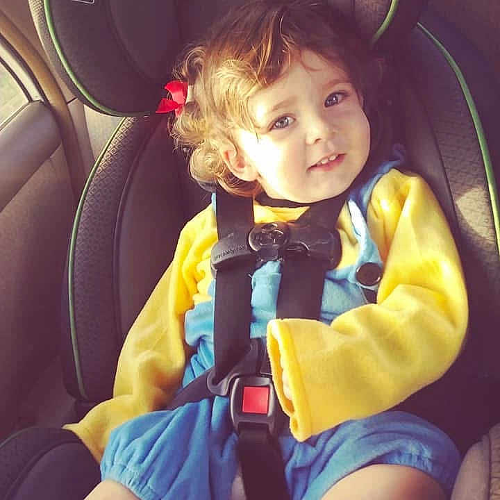 Zayden is registered to the contest to win money with this photo: toddler, child, car_seat, safety_harness, curly_hair, red_bow, yellow_sleeves, blue_overall, smiling, portrait, indoors, car_interior, seatbelt, cute, young_child, happy, person, baby, clothing, seat