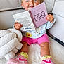 baby, child, girl, pink_bows, book, reading, sneakers, nike, pink_shorts, yellow_socks, blanket, rug, indoor, cute, cozy, smiling, fashion, couch, person, portrait