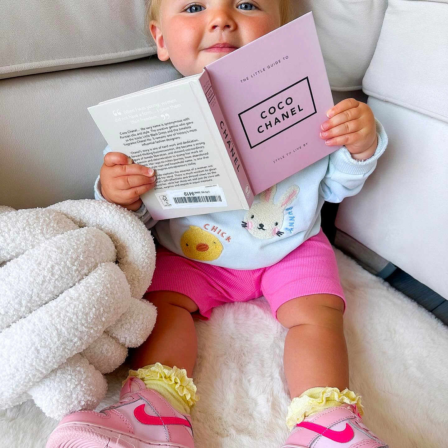 Marlie-Maevela is registered to the contest to win money with this photo: baby, blanket, book, child, couch, cozy, cute, fashion, girl, indoor, nike, person, pink_bows, pink_shorts, portrait, reading, rug, smiling, sneakers, yellow_socks
