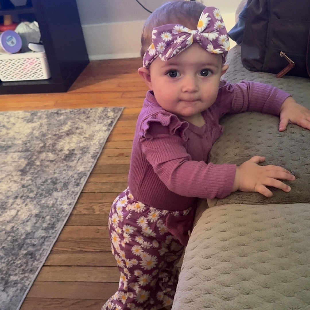 Aubree joined the competition — help win amazing prizes! toddler, child, purple_outfit, headband, daisy_pattern, indoor, living_room, wooden_floor, rug, couch, curious, standing, baby, person, clothing, furniture, home, cute, portrait, smiling