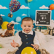 Natanael is registered to the contest to win money with this photo: baby, child, smiling, balloon, blue_sky, clouds, globe, dog_cutout, decor, sign_board, milestone, vest, bow_tie, sitting, indoor, colorful, celebration, toy, portrait, happy