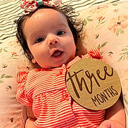 Nicole is registered to the contest to win money with this photo: baby, infant, child, striped_clothing, headband, bow, pillow, floral_pattern, wooden_sign, three_months, cute, smiling, lying_down, closeup, indoors, skin, hand, face, eyes, mouth