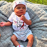 Elias joined the competition — help win amazing prizes! baby, infant, clothing, hat, socks, blanket, outdoor, grass, portrait, cute, soft_texture, sunlight, baseball_theme, headwear, shorts, person, young_child, baby_boy, resting, casual