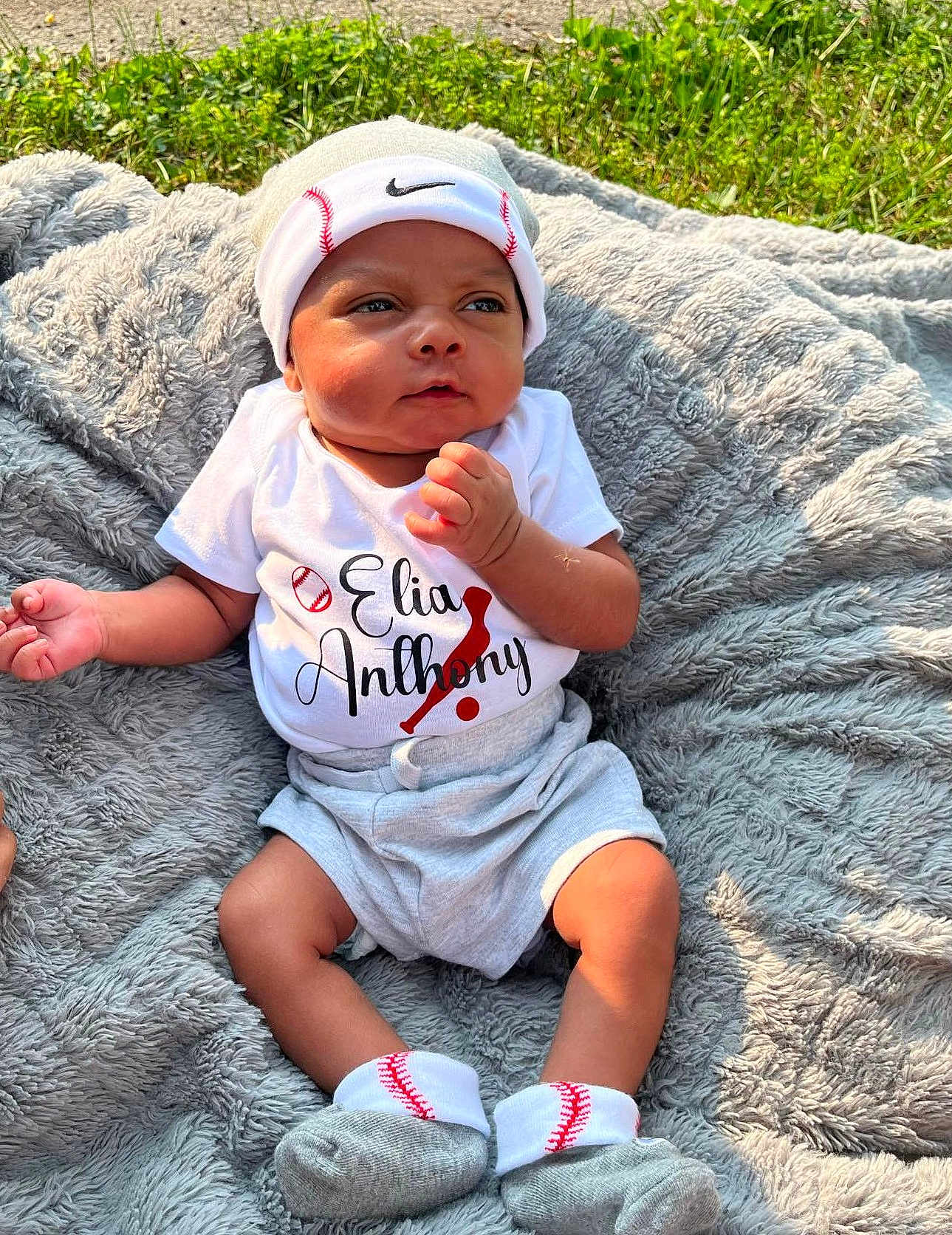 Elias joined the competition — help win amazing prizes! baby, infant, clothing, hat, socks, blanket, outdoor, grass, portrait, cute, soft_texture, sunlight, baseball_theme, headwear, shorts, person, young_child, baby_boy, resting, casual
