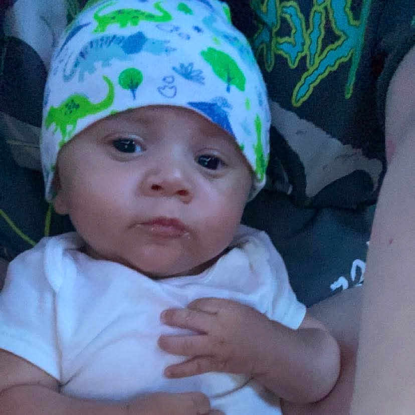 Rhylo is registered to the contest to win money with this photo: baby, babywearing, child, closeup, cute, dinosaur_pattern, expression, eyes, face, hand, hat, headwear, indoor, infant, person, portrait, resting, skin, white_clothing, young