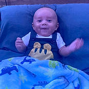 Rhylo is registered to the contest to win money with this photo: baby, smiling, blanket, blue, overalls, animal_print, pillow, wooden_background, happy, infant, child, cute, cozy, clothing, portrait, indoors, face, hands, expression, young