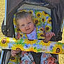 Amelia is registered to the contest to win money with this photo: baby, stroller, sunflower_pattern, cushion, child, outdoor, grass, happy, face, expression, floral_pants, person, seatbelt, sunlight, cheeky, cute, infant, playful, clothing, nature