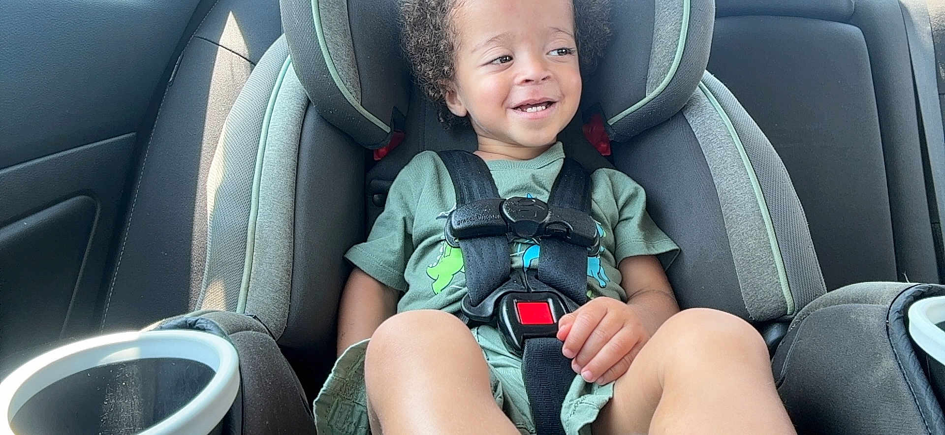 Roman is registered to the contest to win money with this photo: toddler, child, car_seat, smiling, curly_hair, seatbelt, vehicle_interior, safety_harness, green_shirt, happy, portrait, casual_clothing, young_child, daylight, car_door, cup_holder, sitting, cute, baby, kid