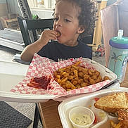 Roman is registered to the contest to win money with this photo: child, curly_hair, eating, fried_food, french_fries, takeout_container, table, sippy_cup, indoor, home, treadmill, chair, food, snack, person, meal, plastic_cup, curly_hair_child, casual, ketchup