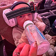 Stetson-Alan is registered to the contest to win money with this photo: baby, bottle, earmuffs, headband, indoor, crowd, seating, event, hand, nail, ring, clothing, warm, feeding, person, audience, soft_lighting, cushion, child, closeup