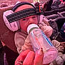 baby, bottle, earmuffs, headband, indoor, crowd, seating, event, hand, nail, ring, clothing, warm, feeding, person, audience, soft_lighting, cushion, child, closeup