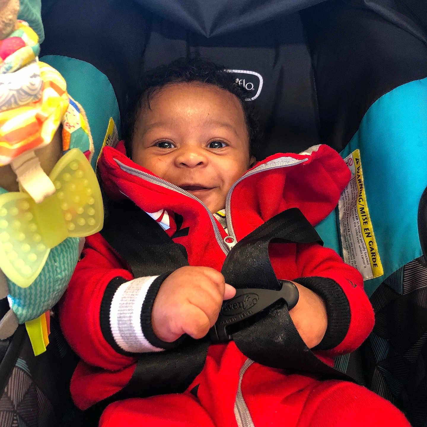 Za is registered to the contest to win money with this photo: baby, child, car_seat, red_clothing, smiling, happy, infant, toy, straps, seatbelt, cute, indoors, portrait, young_child, secure, playful, colorful, close_up, sitting, warm_clothing
