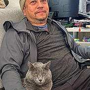 Blakey The Gray Cat is registered to the contest to win money with this photo: man, cat, gray_cat, beanie, glasses, couch, indoor, computer_monitor, desk, relaxed, casual_clothing, sitting, living_room, pet, furniture, window_blinds, cat_tree, technology, home, portrait